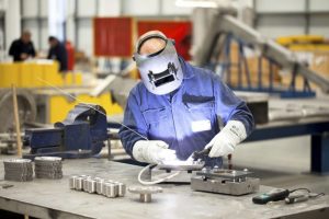 A formal corporate photograph of a engineer at Darchem engineering-Esterline. He is welding mechanical components in the Darchem warehouse and is wearing full personal protective equipment (PPE) including welding head visa, overalls and welding gloves.