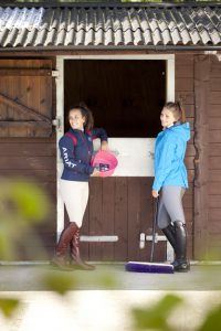 Professional advertising photography shot showing two females in equestrian wear stood outside of a stable with some blurred greenery in the foreground. Shot by professional northeast advertising photographer Cal Carey.
