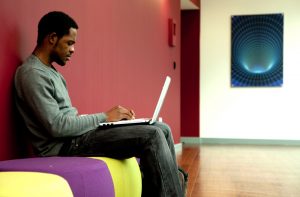 Professional advertising shot showing a young man to the left of the frame sat on colourful seats while on his laptop. Shot by professional northeast advertising photographer Cal Carey.