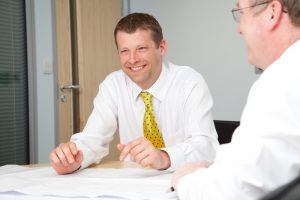 Professional commercial photography shot showing a man sat at a table, grinning with another man on the right of the frame in the foreground and out of focus. Shot by professional northeast commercial photographer Cal Carey.