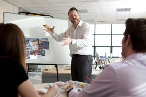 This is a professional photograph of the Velvet Communications brand manager enthusiastically presenting a project, with a whiteboard in the background and his two co-workers giving feedback and reacting to his project. Shot by professional northeast commercial photographer Cal Carey.