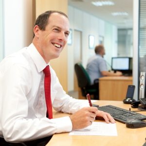 Professional commercial shot showing a mid-shot of a man sat at his desk writing but smiling looking upwards. Shot by professional northeast commercial photographer Cal Carey.