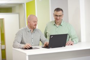 A Professional shot of two workers sat at a desk smiling while working on their computers. Shot by professional northeast commercial photographer Cal Carey.