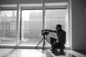 Black and white behind the scenes photography shot showing Cal Carey Photographer kneeling down with his camera on a tripod with a large window behind.