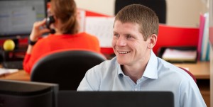 Professional corporate commercial shot of a man sat at his desk, smiling and looking to the left of the frame. Shot by professional northeast commercial and corporate photographer Cal Carey.
