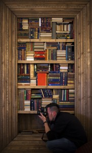 Behind the scenes portrait shot showing Cal Carey the professional advertising photographer crouched down taking a photograph whilst a large bookshelf is behind him. From a professional advertising photography shoot for Barker & Stonehouse.