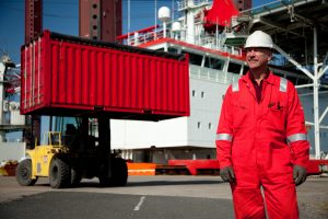 Professional industrial shot showing a worker in red, high-visibility, overalls with a hard hat looking up to the left-hand corner of the frame with a forklift lifting a container up behind him. Shot by professional northeast industrial photographer Cal Carey.