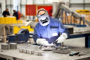 Professional industrial shot showing a worker in blue overalls and a metal visor welding industrial equipment. Shot by professional northeast industrial photographer Cal Carey.