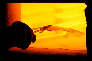 Professional industrial photography shot showing a gloved hand holding some players with a piece of metal in them, going into a melting chamber. Shot by professional northeast industrial photographer Cal Carey.